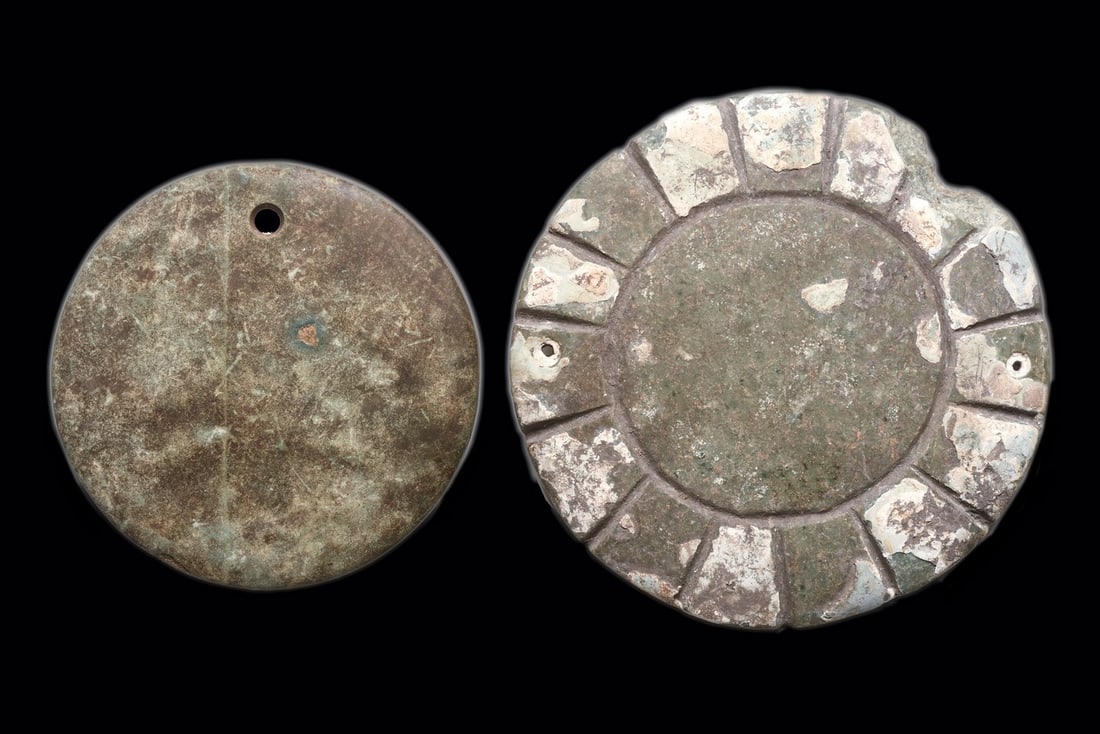 Mesoamerican Circular Stone Pendants (2): West Mexico, ca. 100 BCE – 250 CE. Both are gray green circular pectorals, each with drilled suspension holes. Smaller with a single drill hole and undecorated. Larger wirth remnants of stuccoed
