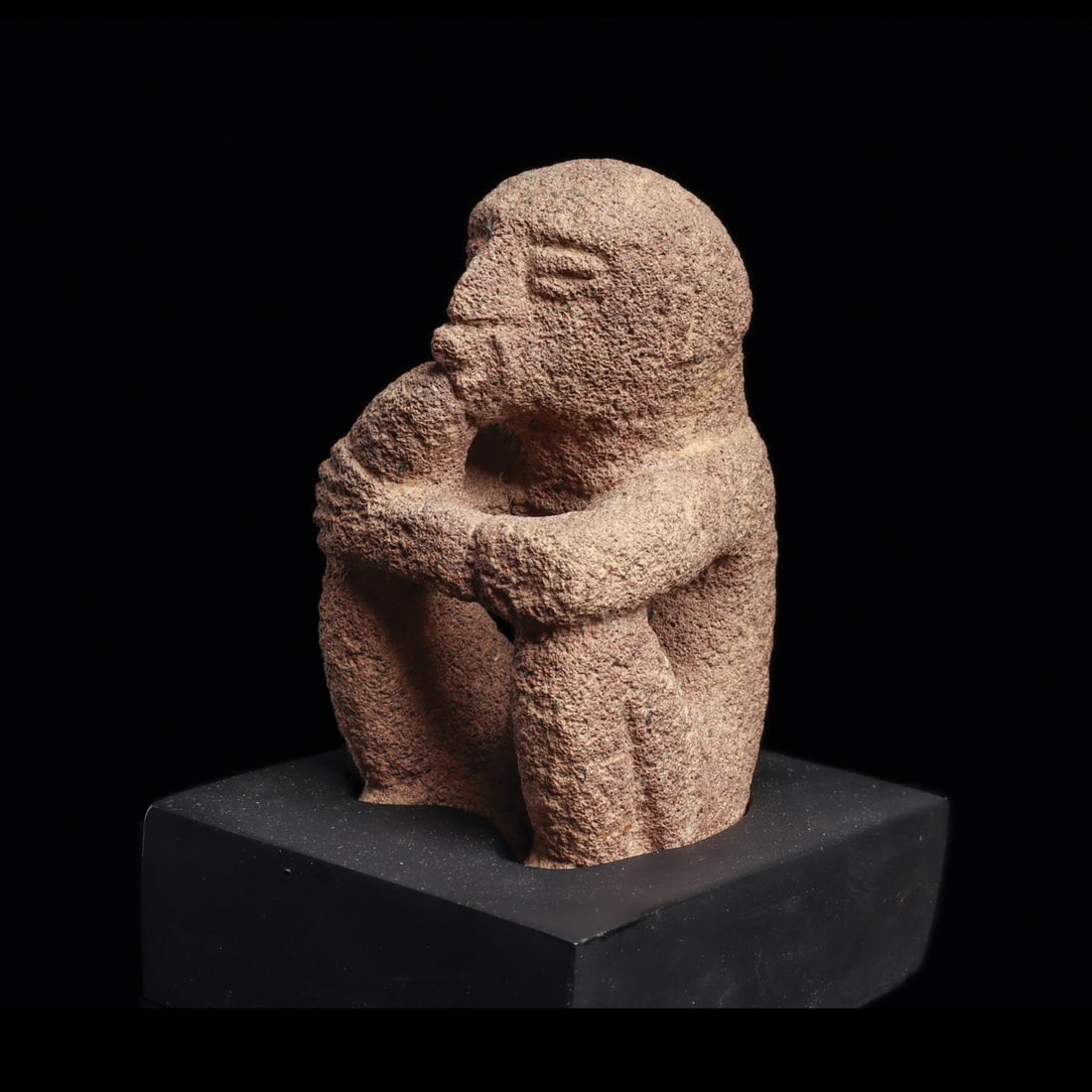 Costa Rican Sukia Figure: Costa Rica, Atlantic Watershed, ca. 10th–14th century CE. Carved gray volcanic stone seated figure, resting both arms on his raised knees, depicted holding his right hand to his mouth, in a smok