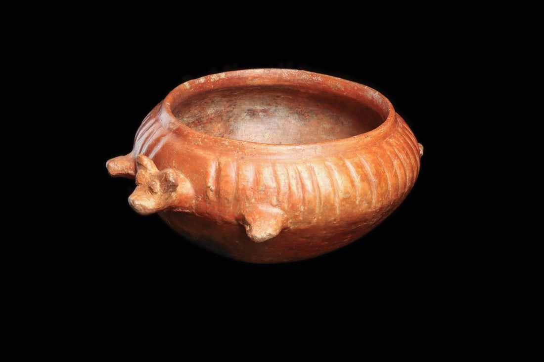 Nayarit Dog Effigy Bowl: Nayarit, ca. 100 BCE – 250 CE. Orangeware pottery effigy bowl depicting a stylized dog having a projecting head, tail and legs, ribbed upper body. Scattered mineral deposits. 7" D (17.78 cm D).