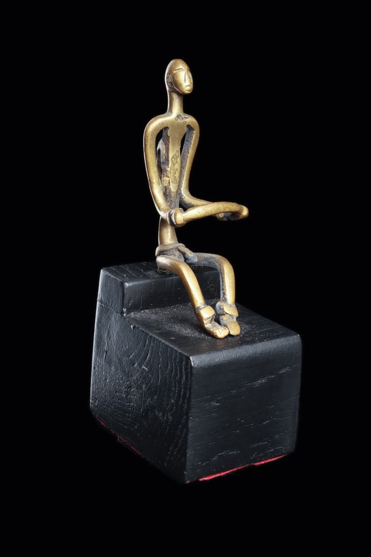 Small Fine West Africa Bronze Figure: Mali. Copper alloy figure of minimalist form, seated with legs slightly curved, and holding a bar in hands. Cuffs adorn wrists and ankles. Face is evoked with simple lines. Exposed phallus. Hanging ho