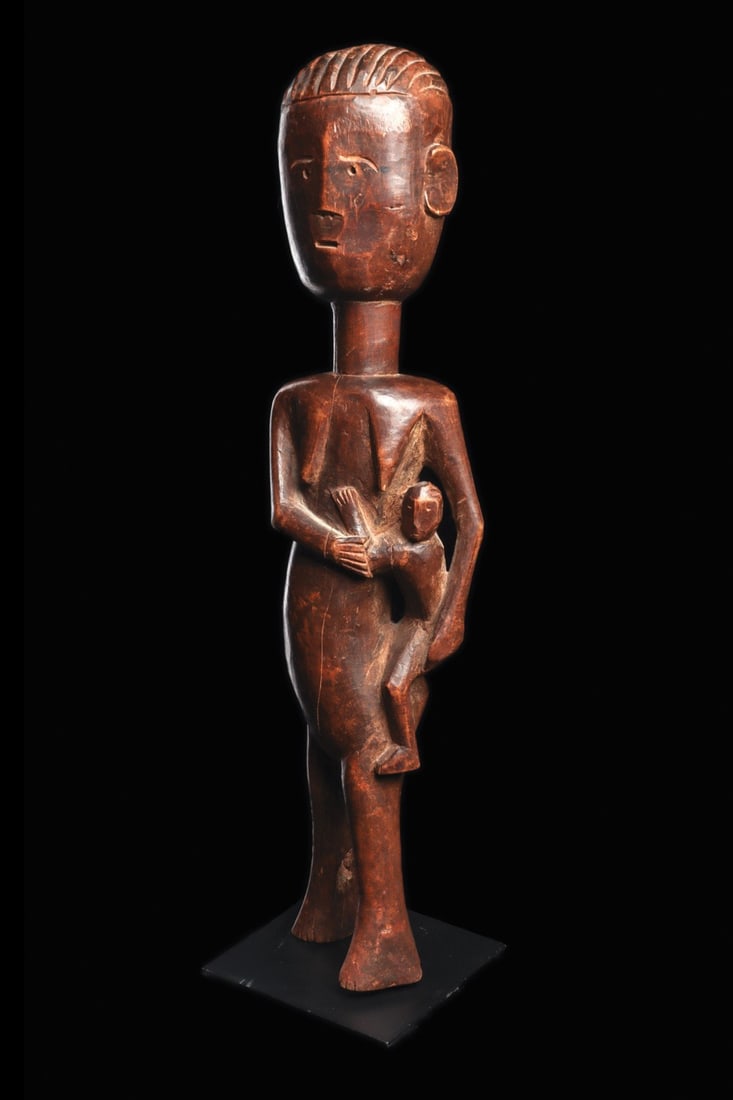 Akan Maternity Figure: Ghana. Charming figure carved of hardwood, the mother stands in twisting position, hoisting small child up along one hip. Red-brown burnished smooth weathered patina. Custom base. 23.75" H (60.33 cm H