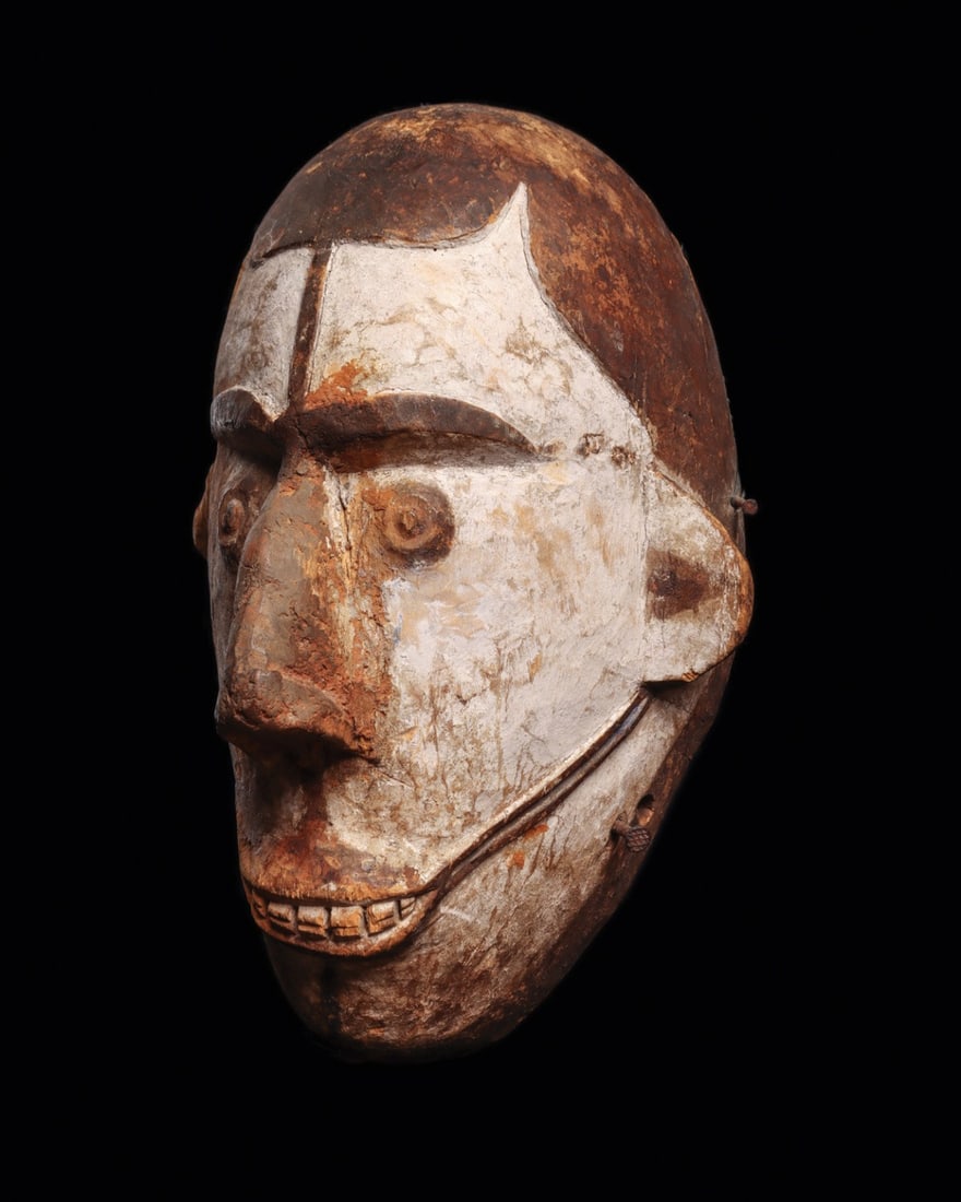 Igbo Crest Mask: Nigeria. Thick ovoid mask of a female spirit having white face with high jutting curved nose, small concentric eyes, and open mouth with defined teeth. The relief line along forehead connecting mouth
