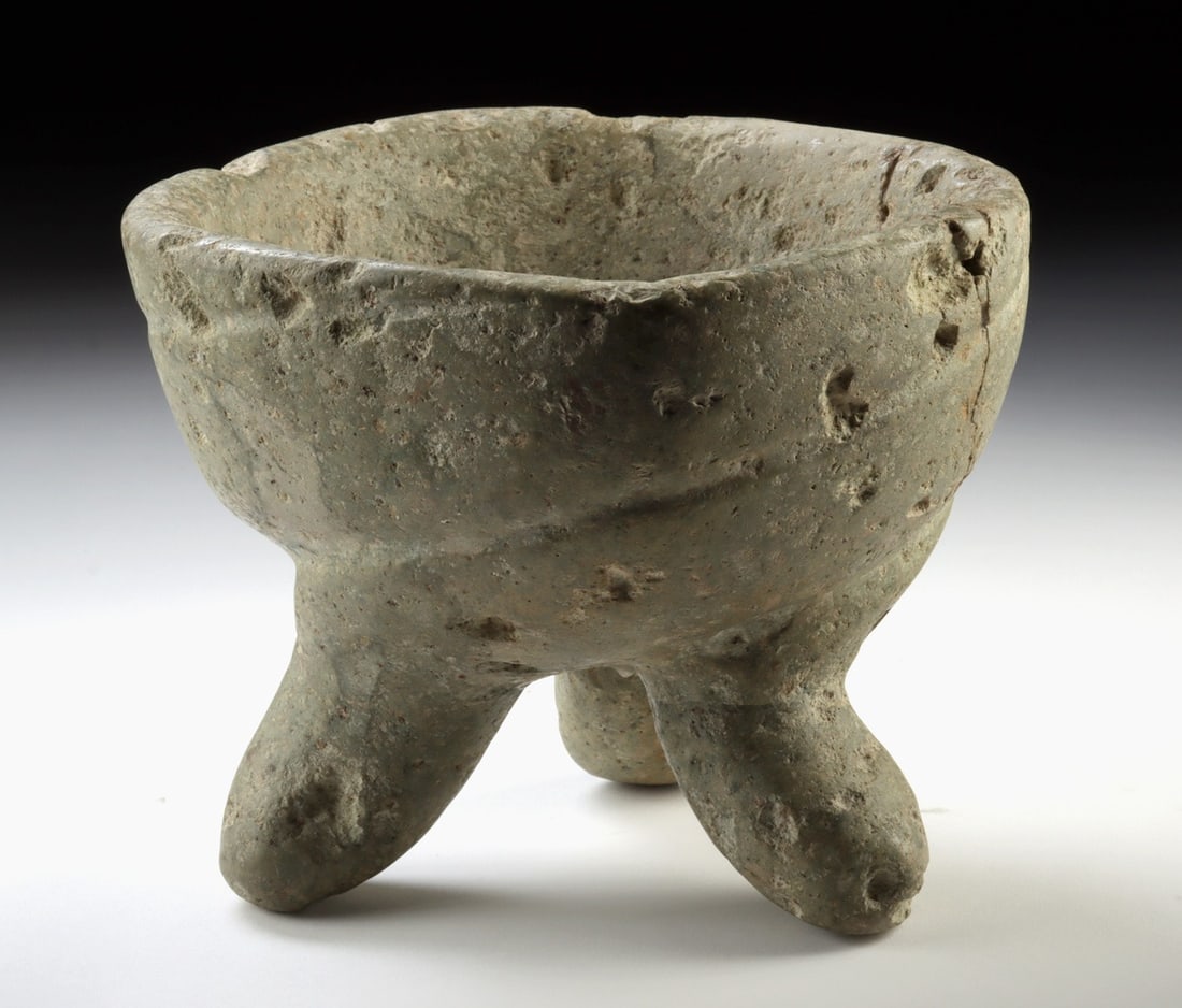 Mixtec Greenstone Tripod Molcajete Mortar: Pre-Columbian, Mesoamerica, Mexico, Mixtec culture, ca. 900 to 1521 CE. A substantial Mixtec stone tripod molcajete carved with a deep circular basin supported by three rounded legs, its surface beari