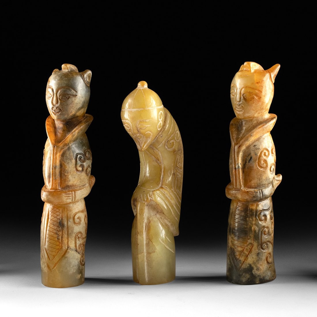 Three Chinese Archaistic Carved Greenstone Figures: East Asia, China, Qing Dynasty to Republic era, ca. early to mid-20th century CE. A group of three standing figures carved from mottled celadon and russet-toned hardstone in an archaistic manner. Two 