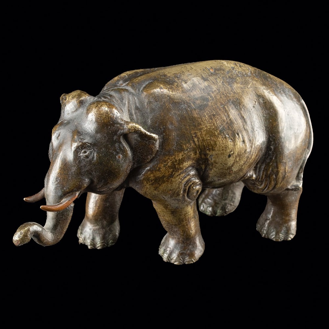 19th C. Japanese Meiji Brass Okimono of an Elephant: East Asia, Japan, Meiji period, ca. 1868–1912 CE. A brass elephant ambles forward on sturdy columnar legs, head lowered and trunk curling inward toward a pair of copper-toned tusks. The modeling