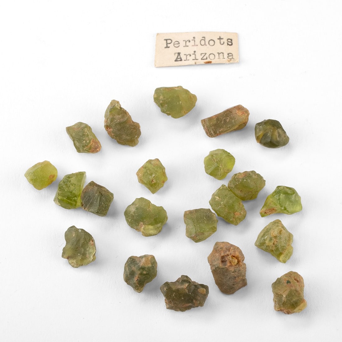 Peridot Gemstones - Raw Olivine Crystals from Arizona: North America, United States, Arizona. A group of twenty-one raw peridot gemstones, 23 grams total, left largely in their natural state, with olive to lime-green color and traces of reddish basaltic m