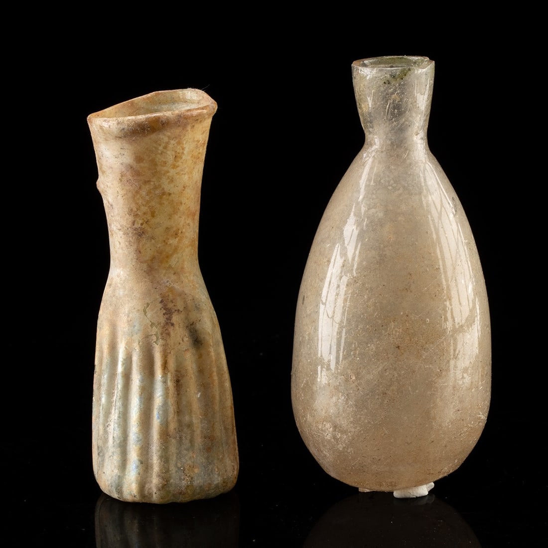 1st-3rd C. Roman Glass Unguentaria - Pair of Ribbed and Piriform Perfume Flasks: Roman, Imperial period, ca. 1–300 CE. A pair of free-blown glass unguentaria vessels. The first is free blown of pale green glass with a slender ribbed body tapering upward to a flared rim, the 