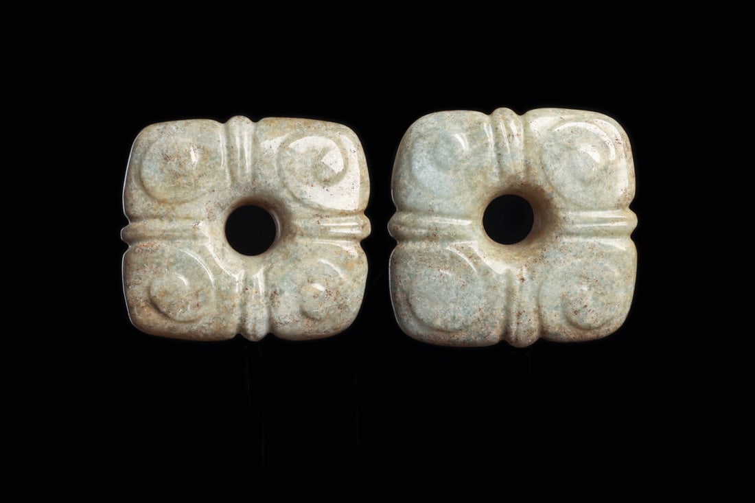 Maya Jade Floral Ear Flare Pair with Spiral Quadrants: Pre-Columbian, Mesoamerica, Maya, ca. 500–800 CE. A matched pair of ear ornaments carved from creamy pale green jade, each a squared quatrefoil whose four petal quadrants carry a raised spiral i