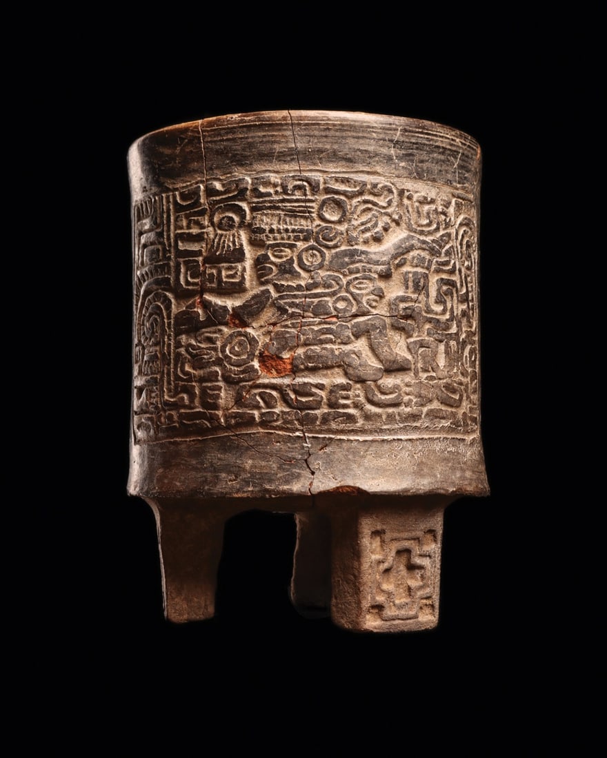 Maya Carved Blackware Tripod Cylinder Vase with Dancing Figure: Pre-Columbian, Mesoamerica, Maya, ca. 500–800 CE. A carved cylinder vase of burnished blackware, its body raised on three integral slab legs, each incised with a stepped concentric cross. Two op