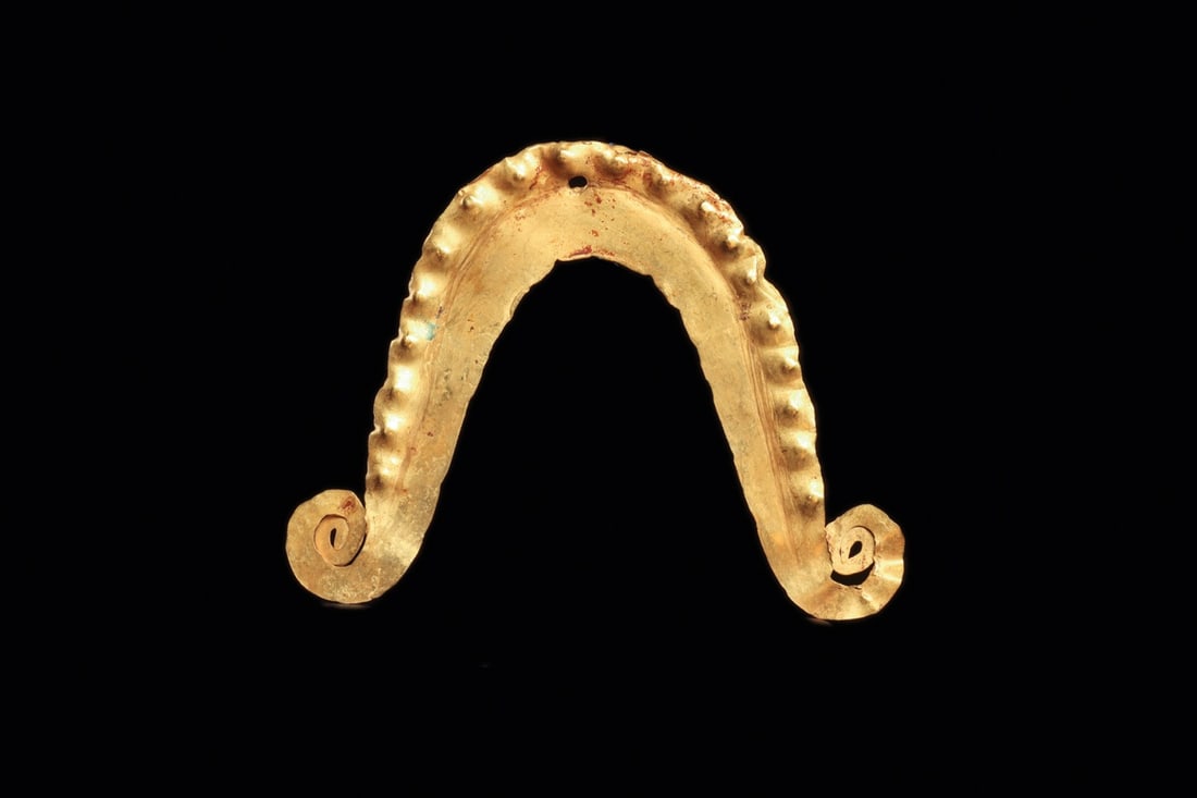 Calima Sheet Gold Mustache Nose Ornament with Spiral Terminals: Pre-Columbian, Colombia, Calima, ca. 500–1000 CE. A crescent of hammered sheet gold bent into a shallow inverted U, its tapering arms curling into tight volutes at each tip. The outer edge carri