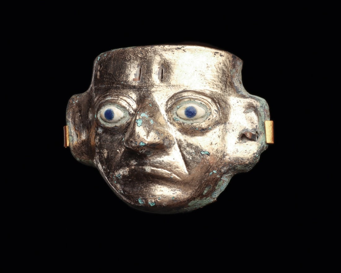 Moche Gilded Maskette with Shell and Lapis Inlay Eyes: Pre-Columbian, Peru, North Coast, Moche, ca. 100–300 CE. A small human face stares out with unnerving directness, its gilded surface still bright across brow and cheek. The maskette was raised a