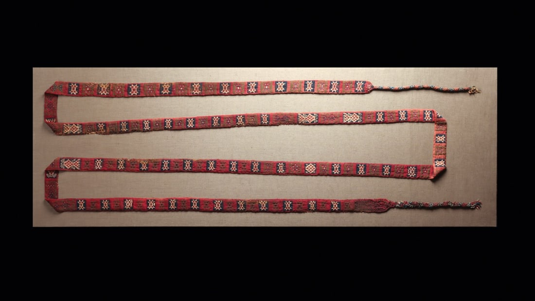 Inca Woven Textile Belt with Geometric Panels, Framed: Pre-Columbian, Peru, Inca Empire, ca. 1250–1450 CE. A long warp-faced band woven in camelid fiber and cotton, its crimson ground divided into a continuous march of framed panels in indigo, cream