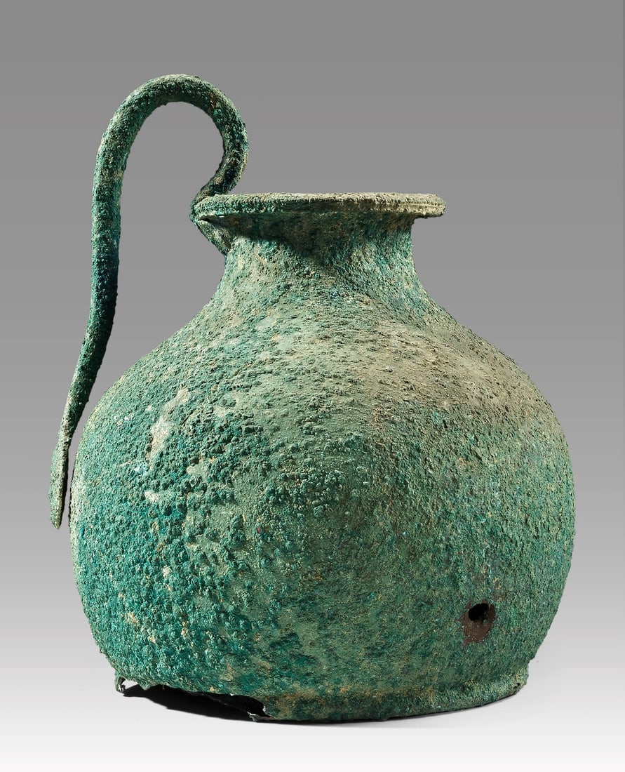 Greek Bronze Oinochoe, Gorgon Handle Roundel: Greece, Classical period, ca. 480–323 BCE. A bronze wine jug whose swelling body rises from a narrow footed base to sloping shoulders, a short neck, and a broad flaring lip finished with a reede