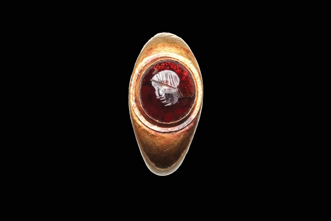 Roman Gold Ring with Garnet Intaglio of a Helmeted Male Head: Roman, Imperial period, ca. 100–300 CE. A gold finger ring of hoop form, the tapered shank swelling to a raised oval bezel that cradles a cabochon garnet. The stone is engraved in intaglio with 