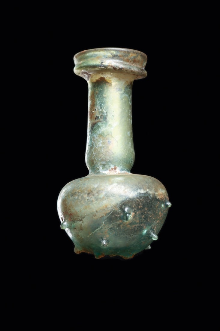 Roman Blue-Green Glass Unguentarium with Pinched Feet: Roman, Imperial period, Eastern Mediterranean, ca. 1–300 CE. A small blown bottle of blue-green metal, its bulbous body rising into a tall cylindrical neck that finishes in a high folded rim. Ar