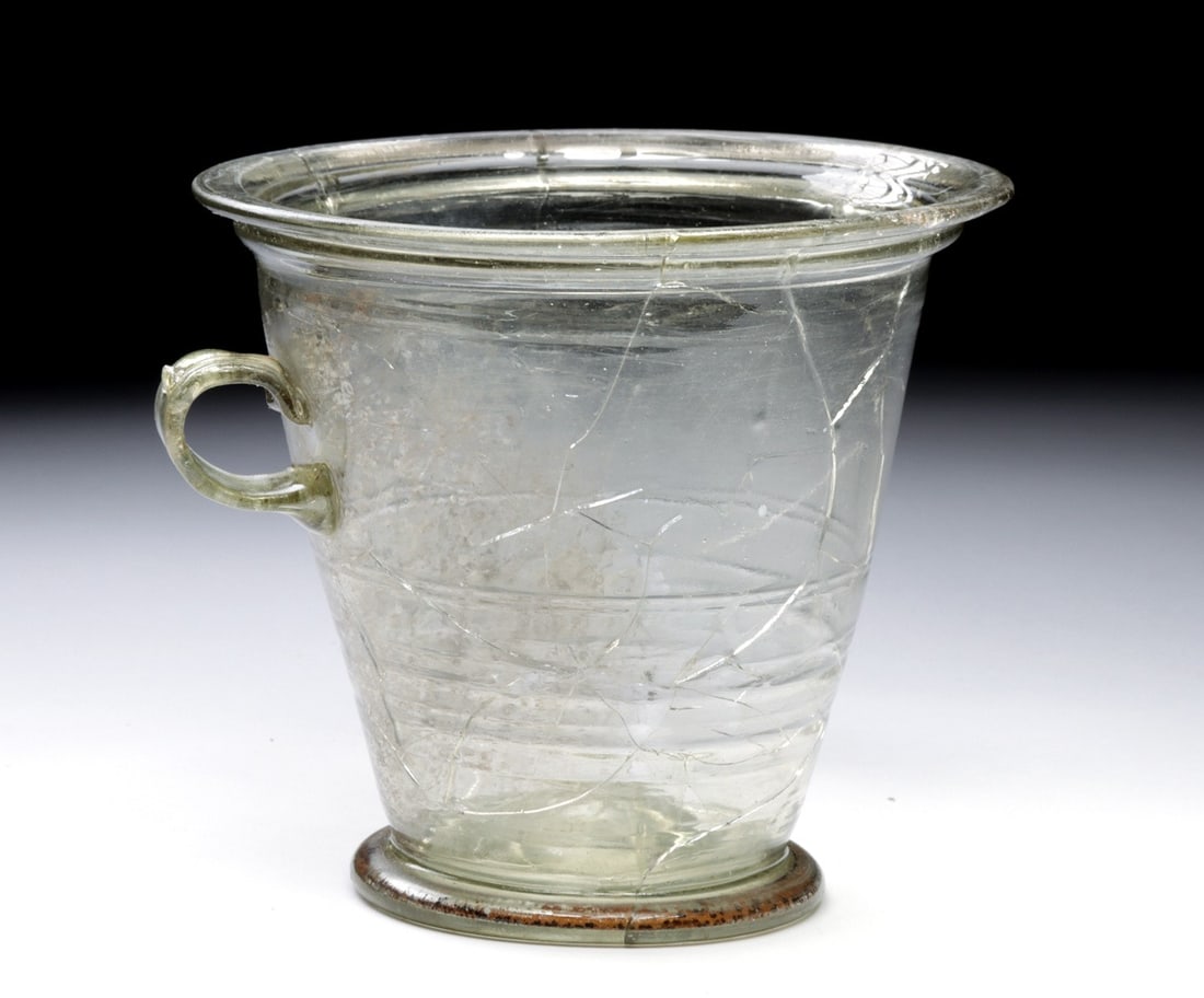 Large Roman Glass Modiolus Cup, Handled Drinking Vessel: Ca. 1–100 CE. Roman, ca. 1st century CE. Drinking cup of pale greenish colorless glass in the modiolus form, a deep, flaring bucket-shaped body set on a thick applied disc foot, with a single sm