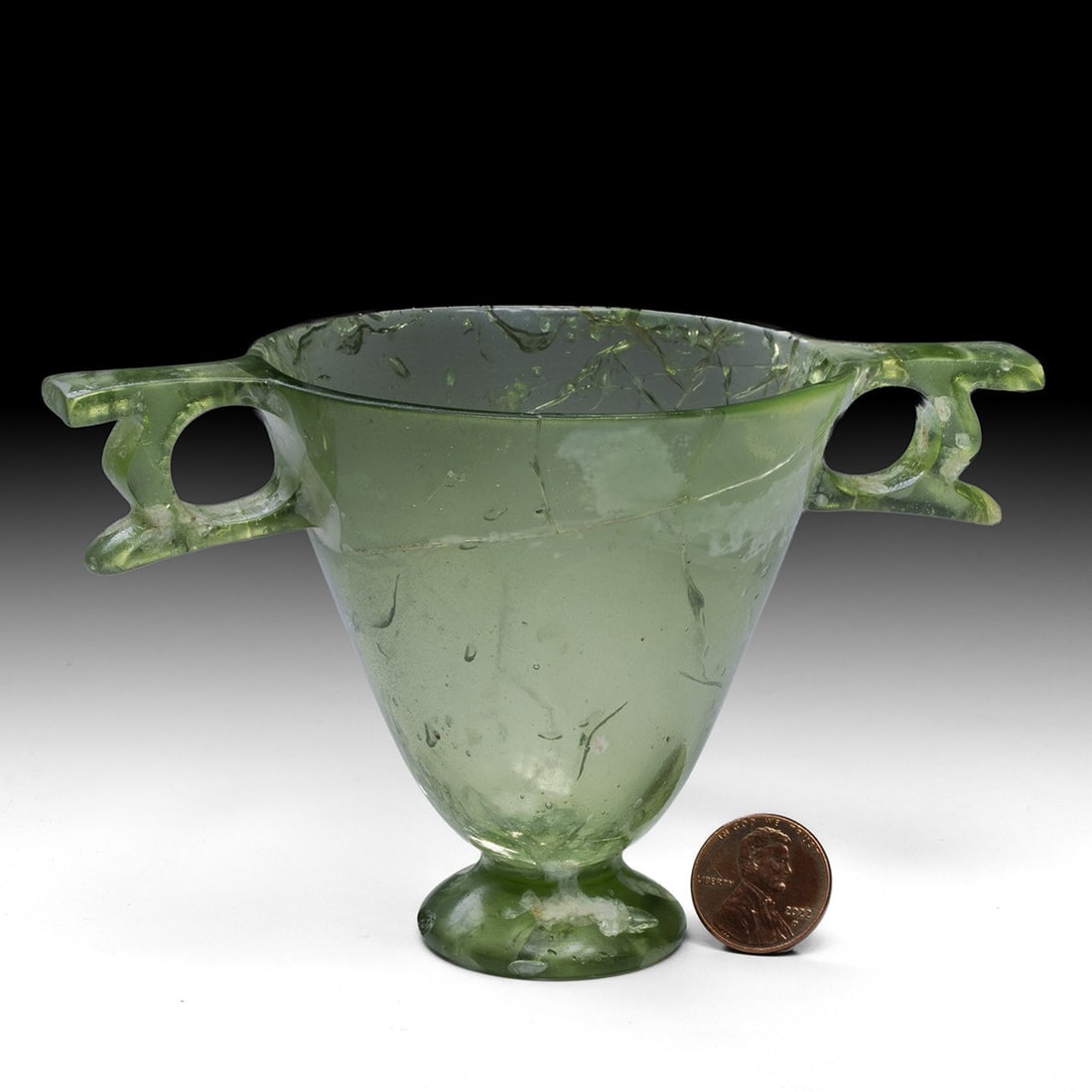 Important Roman Glass Winged-Handle Cup, 1st Century CE: Roman, ca. 0–99 CE. Roman, ca. 1st century CE. Footed cup of pale olive-green glass, with a deep conical body rising from a thick applied pad foot to a wide, plain rim. The handles are two broad