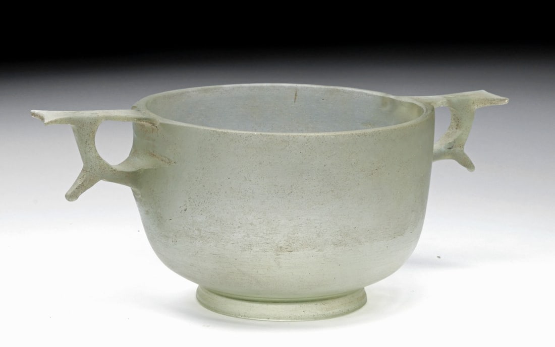 Important Hellenistic Glass Skyphos: Greek, Roman, ca. 250–150 BCE. Glass skyphos of pale olive-green color, cast and cut, with a deep conical body rising from a splayed ring foot to a wide plain rim. Two ring handles are applied to th