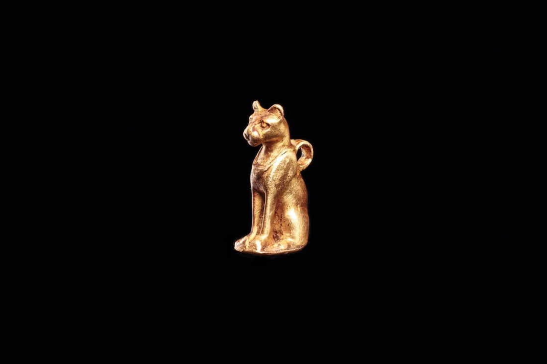 Egyptian Miniature Gold Amulet of Bastet, New Kingdom: Egypt, New Kingdom, 1400 BCE. A miniature solid-cast gold cat sits upright on a thin integral plinth, forelegs straight, haunches gathered, tail curled along one flank. Despite its diminutive scale th