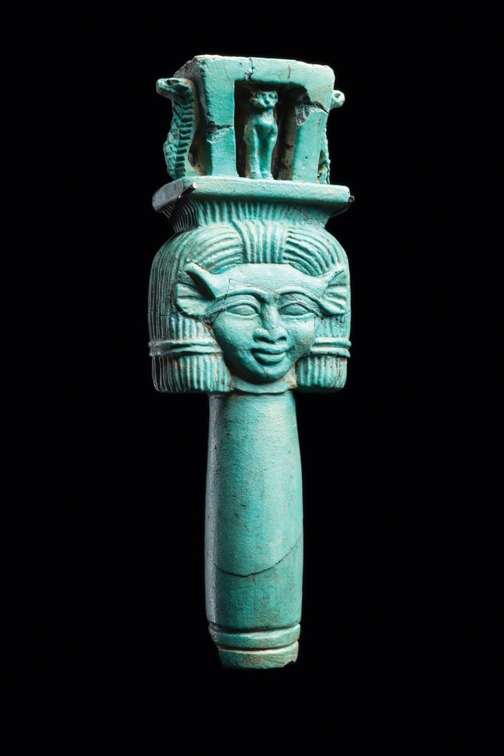 Egyptian Faience Naos Sistrum Handle with Hathor Head and Felines: Egypt, Late Period, 26th Dynasty, ca. 664–525 BCE. A blue-green faience sistrum handle rising into the face of Hathor, cow-eared and framed by the straight striated wig, her mouth set in a faint