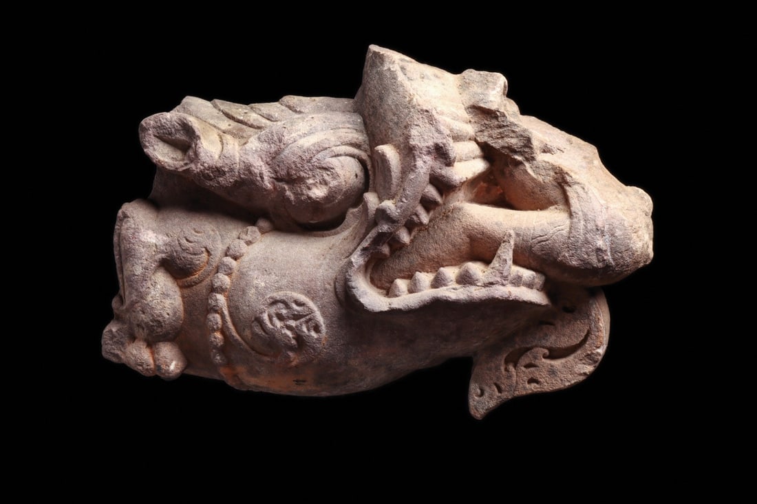 Javanese Sandstone Makara Dragon Head, 10th-16th C.: South East Asia, Indonesia, Java, ca. 900–1600 CE. A red sandstone dragon head carved fully in the round, jaws thrown open around a bristling row of teeth and a curling protruding tongue. The up