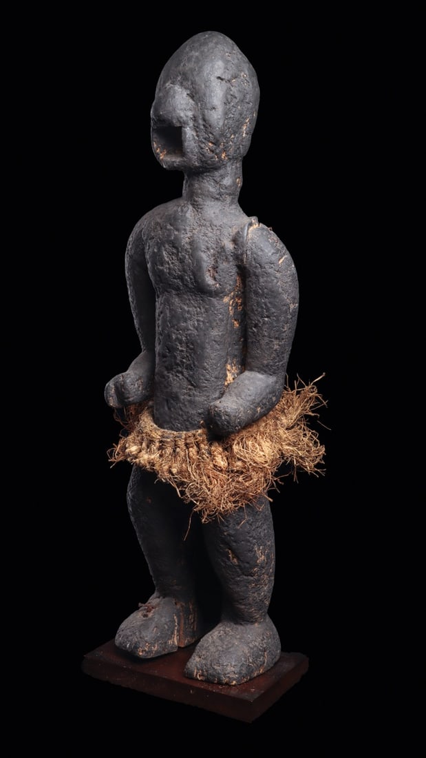 Baule Mbotumbo Monkey Figure with Raffia Skirt, Ivory Coast: West Africa, Ivory Coast, Baule culture, ca. 1900–1960 CE. A hulking guardian carved in wood, part man and part ape, its face nearly swallowed by decades of sacrificial crust. The domed cranium 