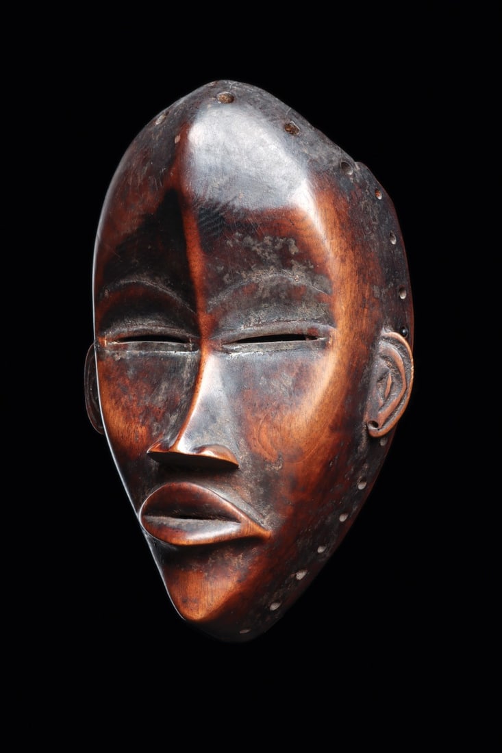 Dan Deangle Mask, Carved Wood Face Mask, Ivory Coast, Early to Mid 20th C.: West Africa, Ivory Coast, Dan peoples, ca. 1900–1960 CE. A serene oval face carved in dense hardwood, its features pared to the Dan ideal of composed beauty. The high domed forehead is divided b