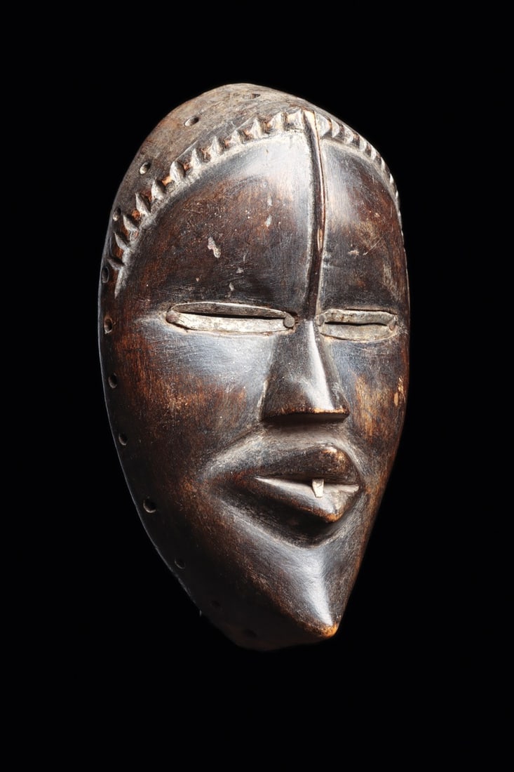 Dan Deangle Spirit Mask with Metal-Lined Slit Eyes, West Africa: West Africa, Ivory Coast, Dan peoples, ca. 1800–1950 CE. A serene oval face carved in wood, its brow divided by a raised central ridge that runs from a notched, saw-toothed hairline to the bridg