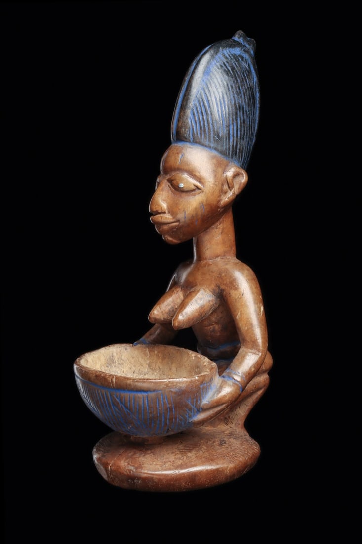 Yoruba Olumeye Bowl Bearer Figure with Indigo Pigment: West Africa, Nigeria, Yoruba, ca. 1800–1920 CE. A kneeling woman rises from an integral circular pedestal, both hands steadying a deep hemispherical bowl incised with radiating hatch panels. Car