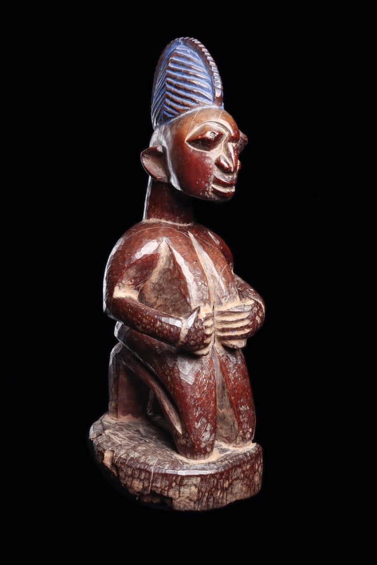Yoruba Kneeling Female Shrine Figure with Indigo Crested Coiffure: West Africa, Nigeria, Yoruba, ca. 1900–1960 CE. A kneeling female devotee carved from a single block of wood, poised on an integral circular base with the tall flat soles of her feet tucked bene