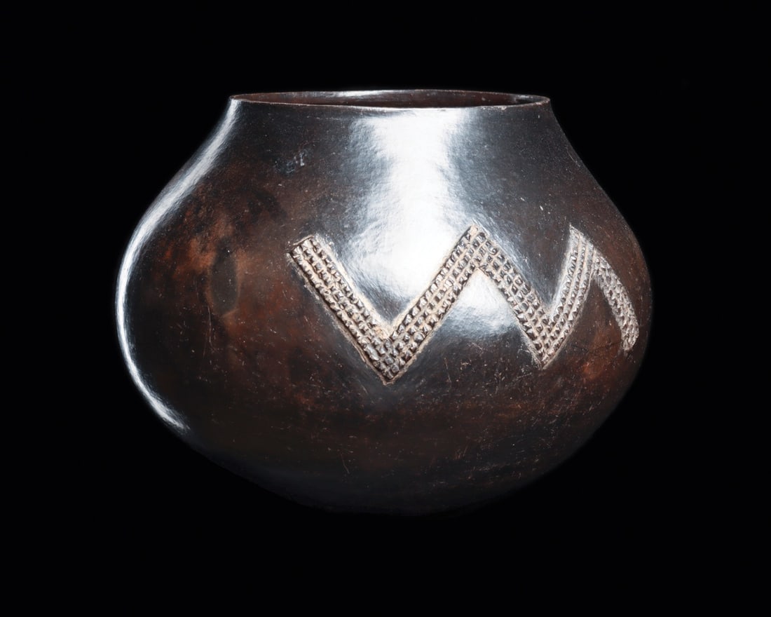 Zulu Ukhamba Beer Vessel with Amasumpa Zigzag Motifs: Southern Africa, South Africa, Zulu, ca. 1800–1950 CE. A generously swelling globular pot, hand-built and burnished to a deep mirror-dark sheen that shifts from graphite black to warm oxblood as
