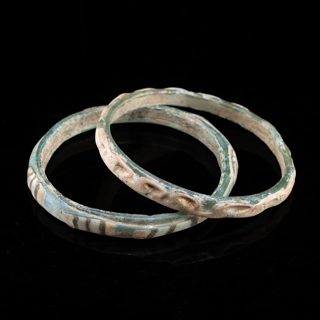 Pair of Byzantine Glass Bracelets w/ Trailing & Stripes: A pair of hand-formed bangles drawn from translucent blue green glass, each closed into a slightly irregular circle that betrays the working of a hot rod around a former. One carries a trailed thread 