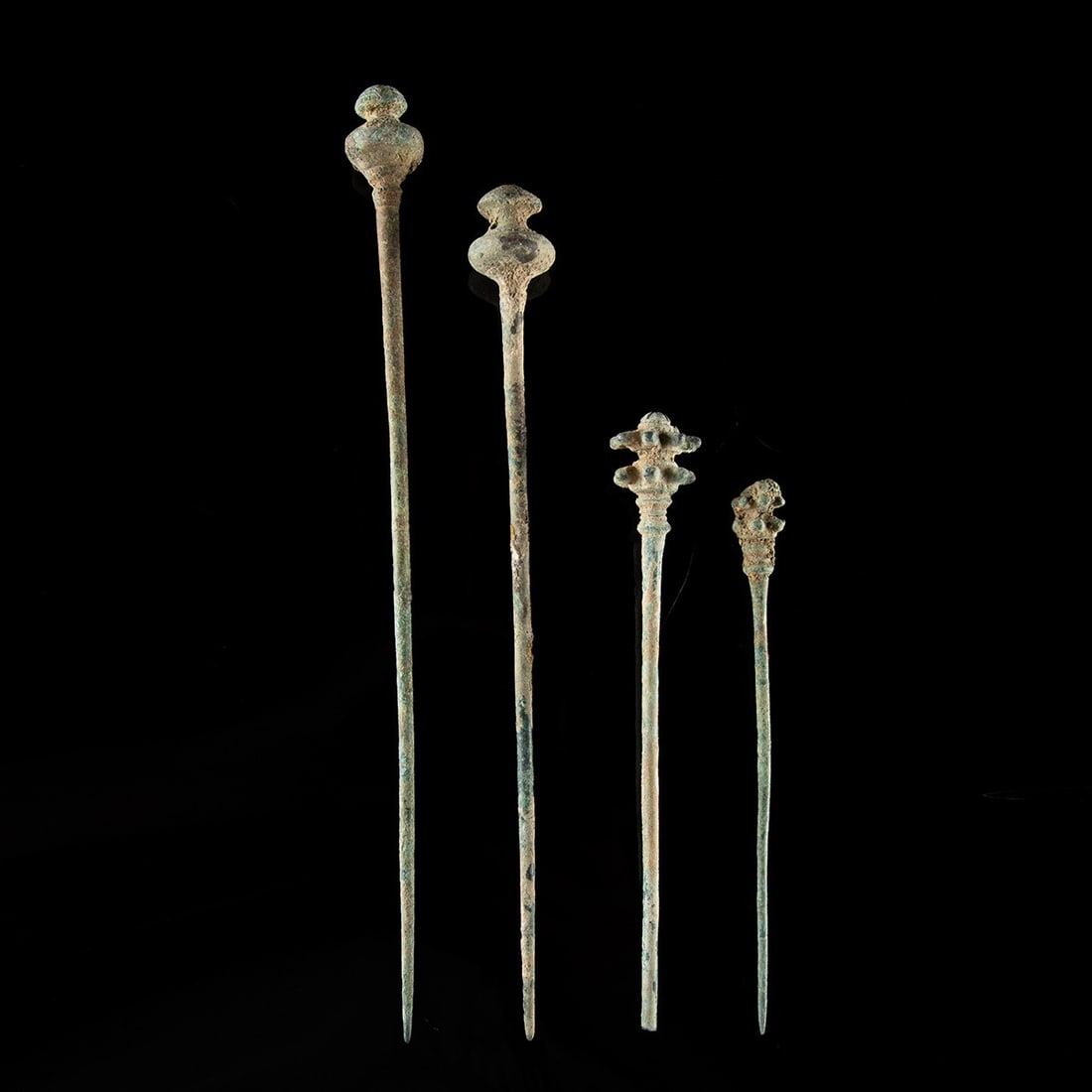 Four Luristan Bronze Garment Pins - Floral & Domed Finials: Northwest Persia, Luristan, ca. 1000–650 BCE. A quartet of long bronze pins from the highland workshops of Luristan, each hand-forged to a slender, tapering shaft and crowned by a distinct decor
