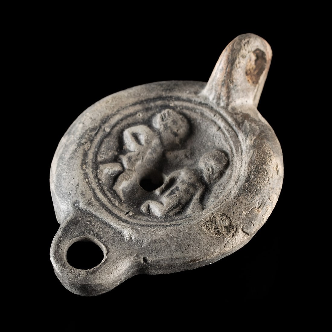 Roman Pottery Oil Lamp w/ Erotic Tondo & Planta Pedis: Roman, Imperial period, ca. 90–140 CE. A greyware oil lamp of classic Imperial form, its rounded nozzle, sloping shoulder, and raised loop handle framing a recessed discus that carries an unabas