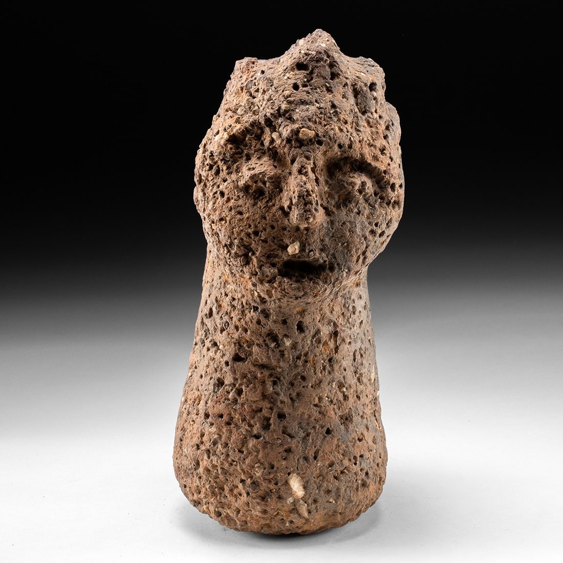Large African Carved Volcanic Stone Head Effigy: Africa, South Sudan, Bongo peoples, ca. 1850–1950 CE. A carved head of red-brown volcanic stone, the highly vesicular surface pitted throughout with open gas cavities and scattered pale inclusio