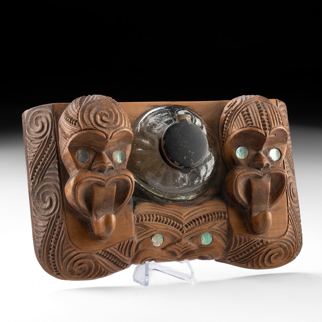 Early 20th C. Maori Carved Wood Inkwell with Paua Shell Eyes: Oceania, New Zealand, Maori culture, ca. 1900–1950 CE. A carved wood desk inkwell holding an antique glass ink jar between tiki figures. A flanking pair of heads each extends a protruding tongue