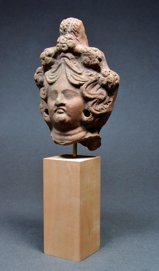 Egyptian Terracotta Female Head - Elaborate Hairdo: Egypt, Early Antoninian Period, ca. 140-150 CE. Sizeable terracotta head of a full-faced woman with fleshy nose, downturned mouth, double chin and Venus folds along the heavy neck. The modeling of the