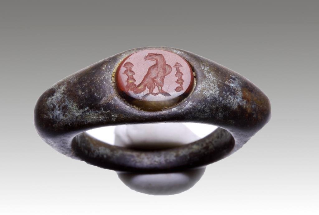 Roman Bronze / Carnelian Ring: Rome, ca. 1st - 5th century CE. A cast bronze ring with carnelian inset. Inset is carved with an eagle standing between two poles/trees. Red carnelian with white stripe across the bottom. Just under 3