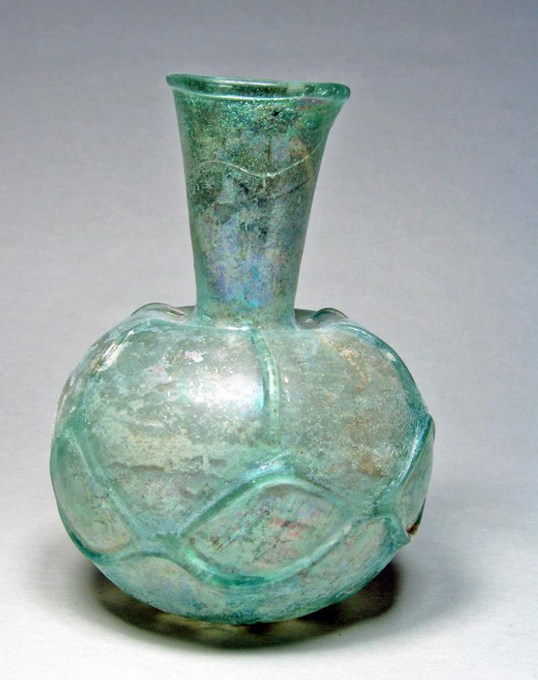 Roman Glass Bottle - Faceted Detailing: Rome, ca. 2nd-3rd Century CE. A fine glass bottle of faceted globular body and funnel shaped neck. Tooled ribs radiating on the shoulder and on the lower body to join a chain pattern around the belly.