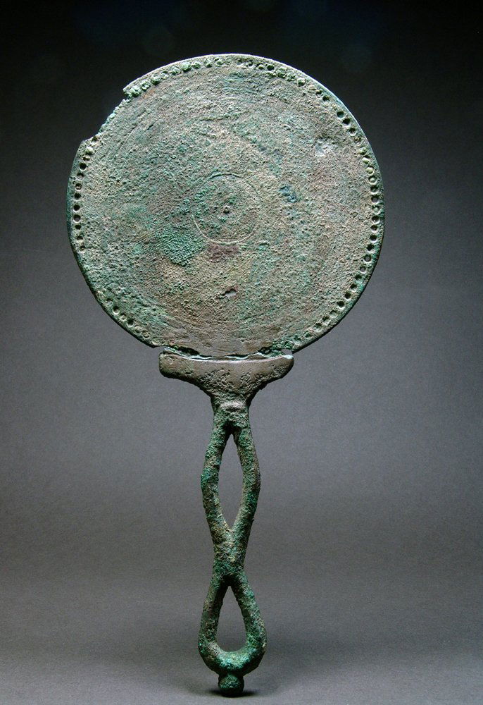 Roman Bronze Hand-Held Mirror: Rome, ca. 2nd / 3rd Century CE. Imperial bronze mirror with a riveted rim and fine concentric lines. The other side would have been burnished for reflection. A lotus blossom at the join of the slung h
