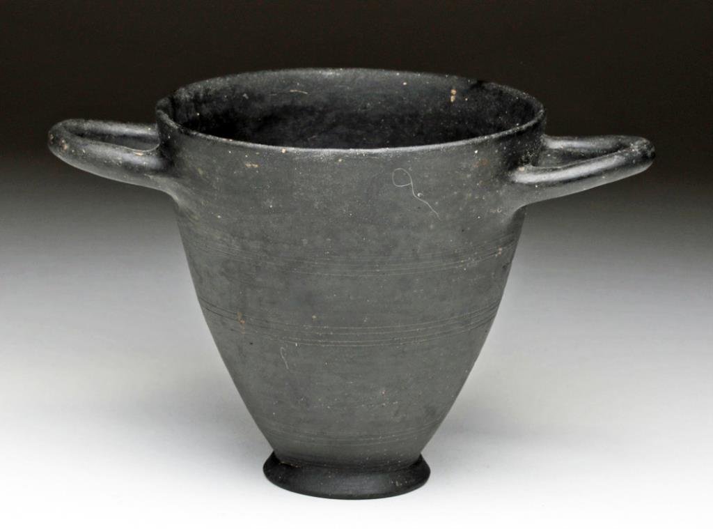 Etruscan Bucchero Pottery Skyphos: Northern Italy, Ca 5th century BCE. Pottery skyphos, or wine cup, in Bucchero blackware, borrowing the style directly from the Greeks of the period. With flared base and twin horizontal handles just b