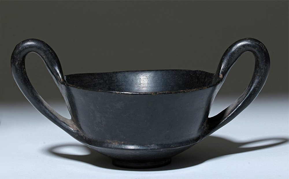 Etruscan Bucchero Kantharos: Central Italy, Etruria, ca. 550 BCE. This is an open burnished blackware vessel on a ring base with high twin strap handles at either side of the rim and a decorative band around the waist. Etruscan b
