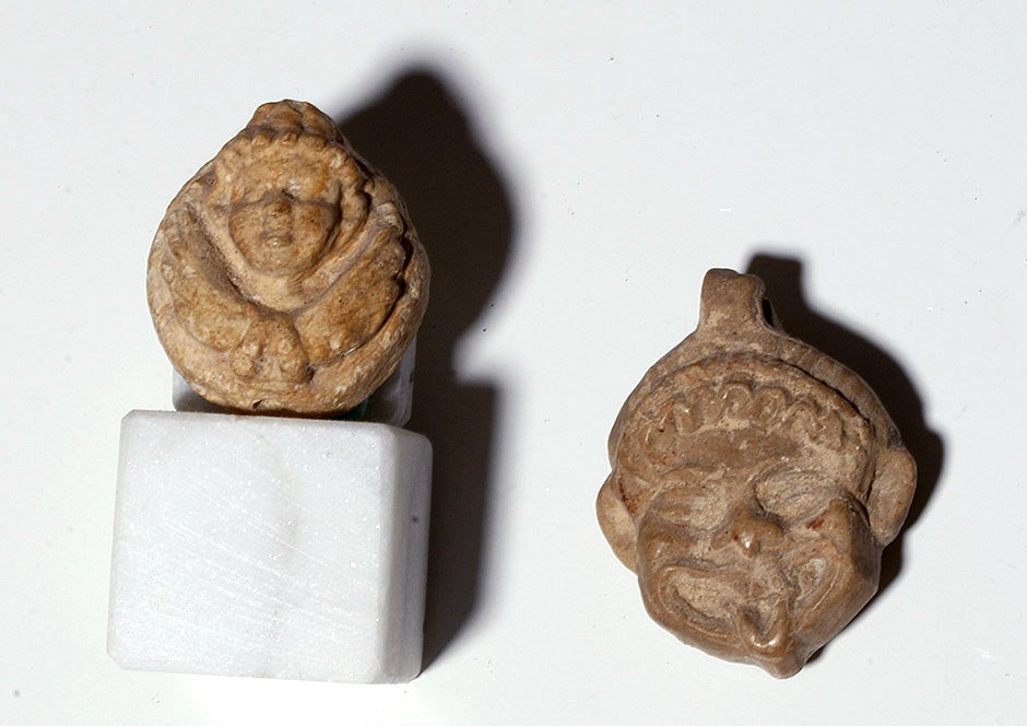 Pair of Roman Stone Face Carvings: Rome, ca. 200 BCE to 300 CE. These are two small pottery carvings of human faces; one, of a slightly grotesque figure with curly hair and his tongue extended, and the other of a more dignified face ab
