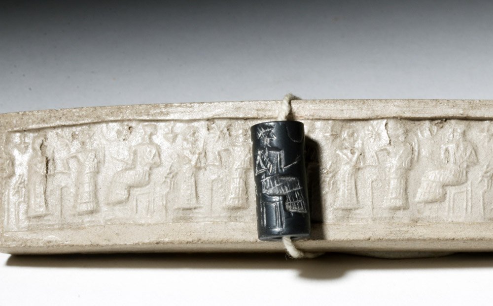 Museum-Exhibited Babylonian Steatite Cylinder Seal: Ancient Sumeria/Mesopotamia, ca. 11th century BCE. A small steatite cylinder seal, its surface engraved in intaglio with a pair of figures standing before a lord or deity, stars above. When rolled in 