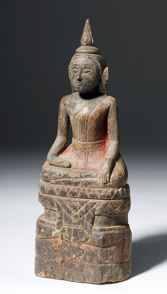 Late 19th C. Laotian Painted Wood Devotional Buddha: Southeast Asia, Laos, ca. 1900 CE. A carved wooden Buddha with some remaining cinnabar pigment and a serene, simple face. He sits atop a tall throne with a lotus design on its upper third. This Buddha