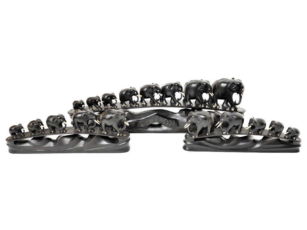 A LOT OF THREE EBONIZED WOOD ELEPHANT CARVINGS, Mi: A LOT OF THREE EBONIZED WOOD ELEPHANT CARVINGS, Mid-Twentieth Century, Ceylon (now known as Sri Lanka) of several elephants walking in a line across a bridge' with ivory tusks; the largest carved ""Ce
