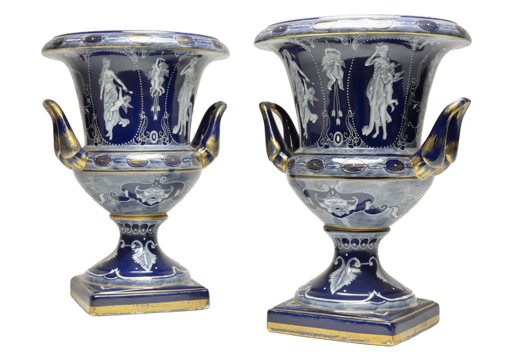 A PAIR OF MINTON STYLE CAMPAGNA TWO HANDLE PORCELA: A PAIR OF MINTON STYLE CAMPAGNA TWO HANDLE PORCELAIN URNS19th Century Each vase with everted lip features a Neoclassical style register of cobalt blue ground decorated in white with Classical motifs o