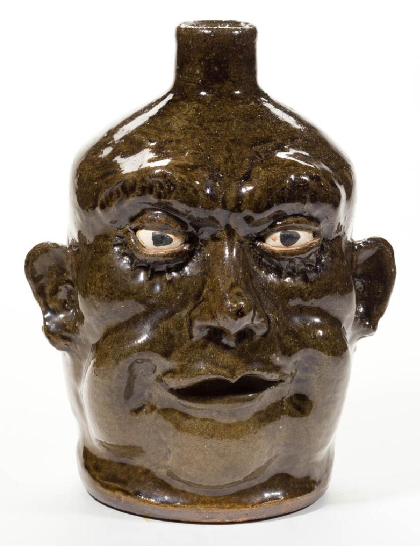 SIGNED "LANIER MEADERS" GEORGIA STONEWARE FACE JUG: SIGNED "LANIER MEADERS" GEORGIA STONEWARE FACE JUG, overall dark green alkaline glaze, with inset ceramic eyes, applied ears, nose, and eyebrows, and white clay teeth. Incised "Lanier / Meaders" on ba