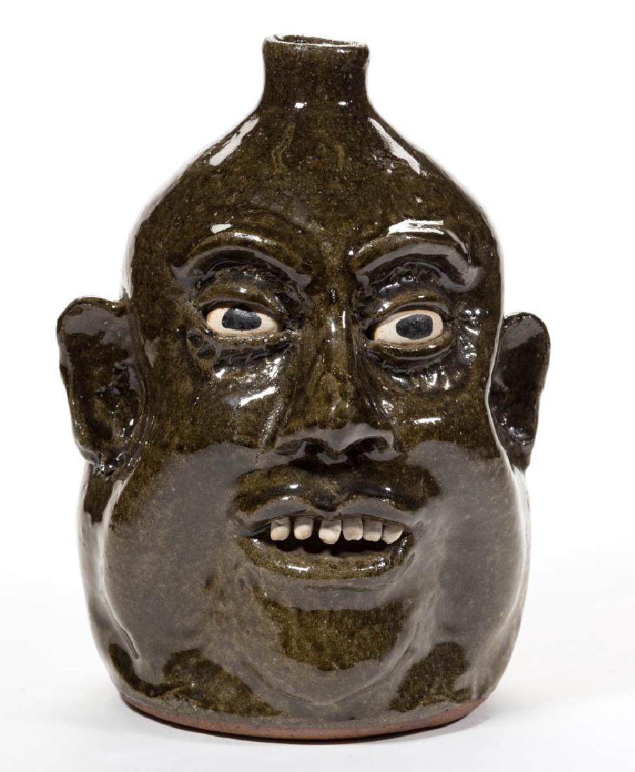 SIGNED "LANIER MEADERS" GEORGIA STONEWARE FACE JUG: SIGNED "LANIER MEADERS" GEORGIA STONEWARE FACE JUG, overall dark green alkaline glaze, with inset ceramic eyes, applied ears, nose, and eyebrows, and white clay teeth. Incised "Lanier / Meaders" on ba