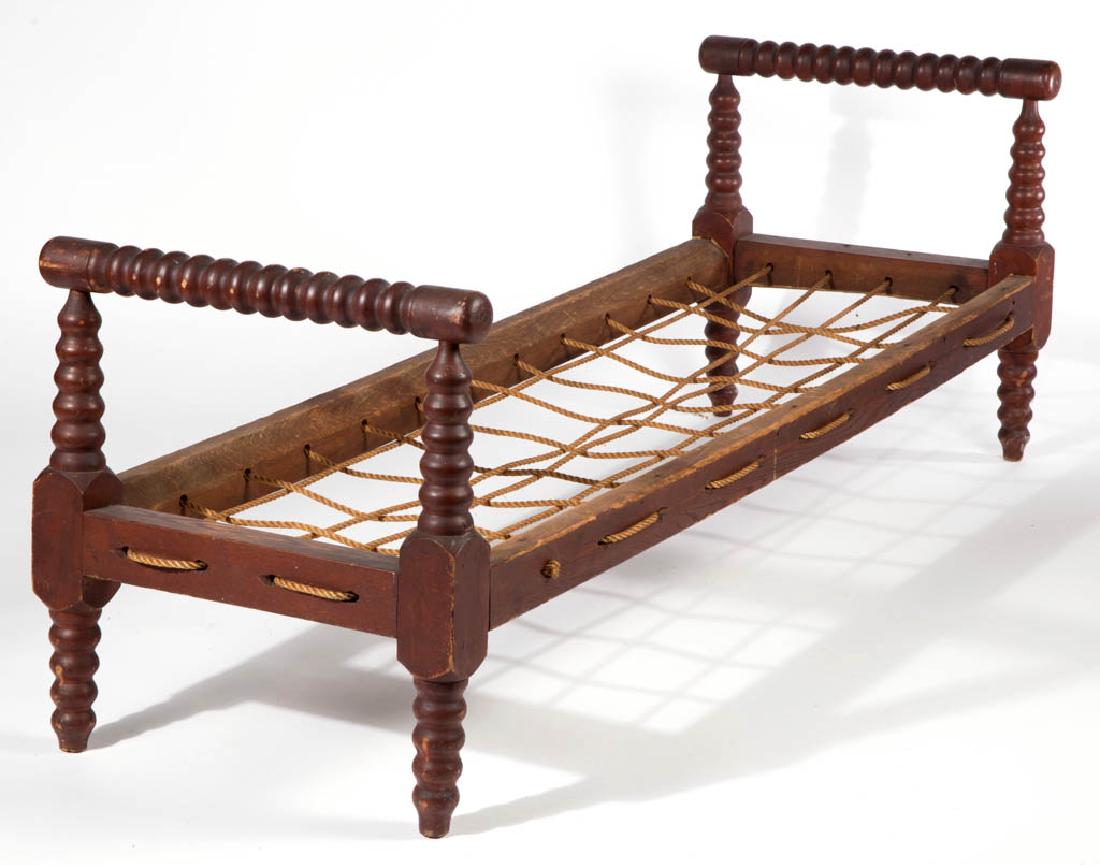 AMERICAN PAINTED OAK AND MAPLE HIRED-MAN'S ROPE BED: AMERICAN PAINTED OAK AND MAPLE HIRED-MAN'S ROPE BED, modified spool turnings, original red-painted surface. Mid 19th century. 25" H, 23 3/4" W, 68 1/4" D.Excellent overall condition with minor expecte